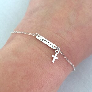 May include: Silver chain bracelet with a small cross charm and a rectangular tag with the name "CECILIA" engraved on it.