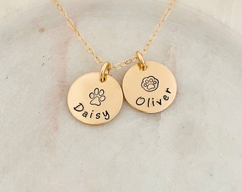 May include: Gold necklace with two round charms. One charm says "Daisy" with a paw print and the other charm says "Oliver" with a paw print.