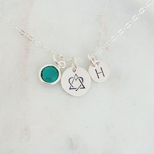 Puede incluir: Un collar de plata con tres colgantes: una piedra preciosa verde, un colgante con un corazón y un triángulo, y un colgante con la letra H.