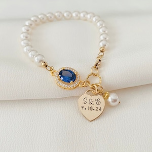 Op de afbeelding: Een gouden armband met een blauwe edelsteen en een hartvormige hanger gegraveerd met "S & S 9.10.24". De armband is gemaakt van witte parels.