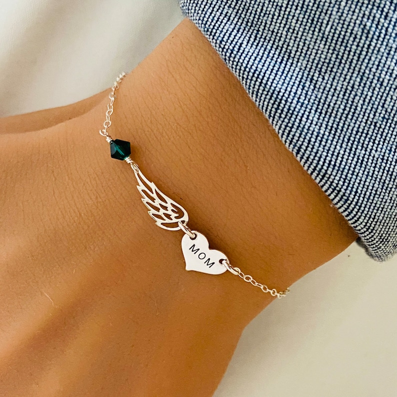 K&ouml;nnte beinhalten: Ein silbernes Armband mit einem Fl&uuml;gelanh&auml;nger, einem Herzanh&auml;nger mit der Gravur "MOM" und einem dunkelgr&uuml;nen Kristall. Das Armband ist durch eine feine Kette verbunden.