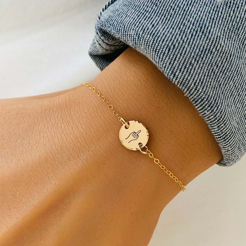 Puede incluir: Un brazalete de cadena dorada con un dije redondo que presenta una mano haciendo el dedo medio.