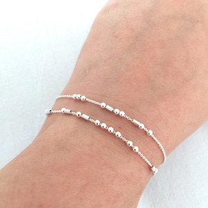 May include: Two silver chain bracelets with small round beads and a few cylindrical beads. The bracelets are layered on a person's wrist.