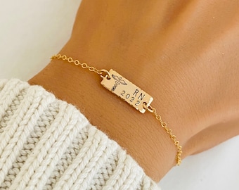 Puede incluir: Pulsera de oro con un dije rectangular que dice "RN 2022" y tiene un símbolo médico grabado.