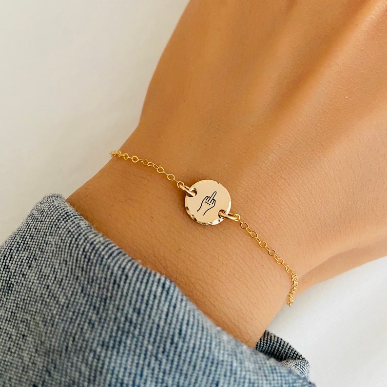 Puede incluir: Un brazalete de cadena dorada con un dije redondo que muestra un dedo medio.