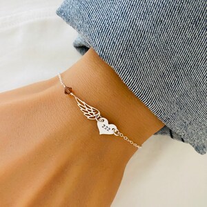 K&ouml;nnte beinhalten: Ein silbernes Armband mit einem Fl&uuml;gel-Anh&auml;nger, einem Herz-Anh&auml;nger mit der Zahl "222" und einer kleinen braunen Kristallperle. Das Armband wird an einem Handgelenk vor einem Jeanshintergrund gezeigt.