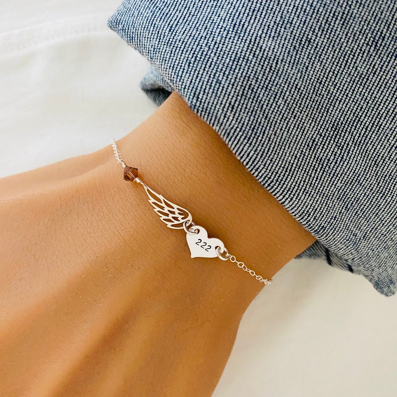 K&ouml;nnte beinhalten: Ein silbernes Armband mit einem Fl&uuml;gel-Anh&auml;nger, einem Herz-Anh&auml;nger mit der Zahl "222" und einer kleinen braunen Kristallperle. Das Armband wird an einem Handgelenk vor einem Jeanshintergrund gezeigt.