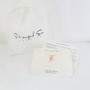 May include: Gold necklace with a small angel wing charm and a round initial charm. The necklace is displayed on a white card with the text "Stamped Eve Jewelry" and "Simple, Modern & Delicate" along with the website address "StampedEve.etsy.com".