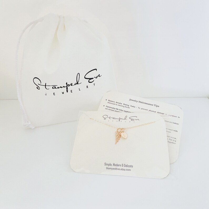 May include: Gold necklace with a small angel wing charm and a round initial charm. The necklace is displayed on a white card with the text "Stamped Eve Jewelry" and "Simple, Modern & Delicate" along with the website address "StampedEve.etsy.com".