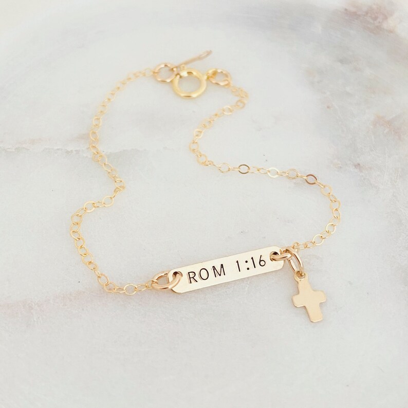 May include: Gold chain bracelet with a small cross charm and a rectangular tag engraved with "ROM 1:16".