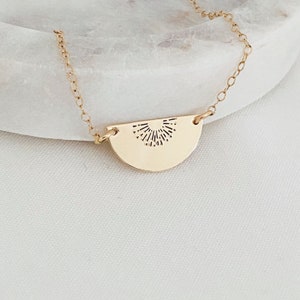 May include: A gold necklace with a delicate chain and a half-circle pendant featuring a sunburst design.