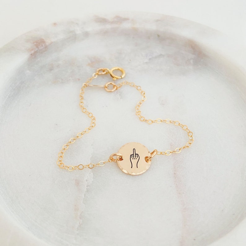 Puede incluir: Pulsera de cadena dorada con un peque&ntilde;o dije redondo que muestra un gesto de dedo medio.