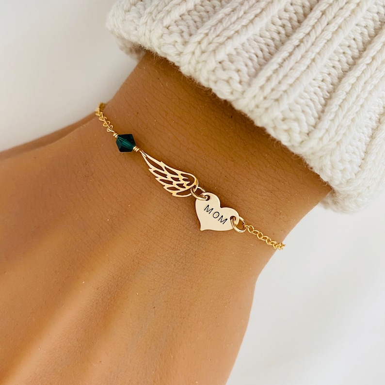 K&ouml;nnte beinhalten: Ein goldfarbenes Armband mit einer filigranen Kette. Es hat einen Herzanh&auml;nger mit dem Wort "MOM", einen Fl&uuml;gelanh&auml;nger und eine dunkelgr&uuml;ne Kristallperle. Das Armband wird am Handgelenk getragen.