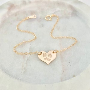 May include: A gold heart-shaped charm bracelet with a delicate chain. The charm features an engraved design of two footprints.