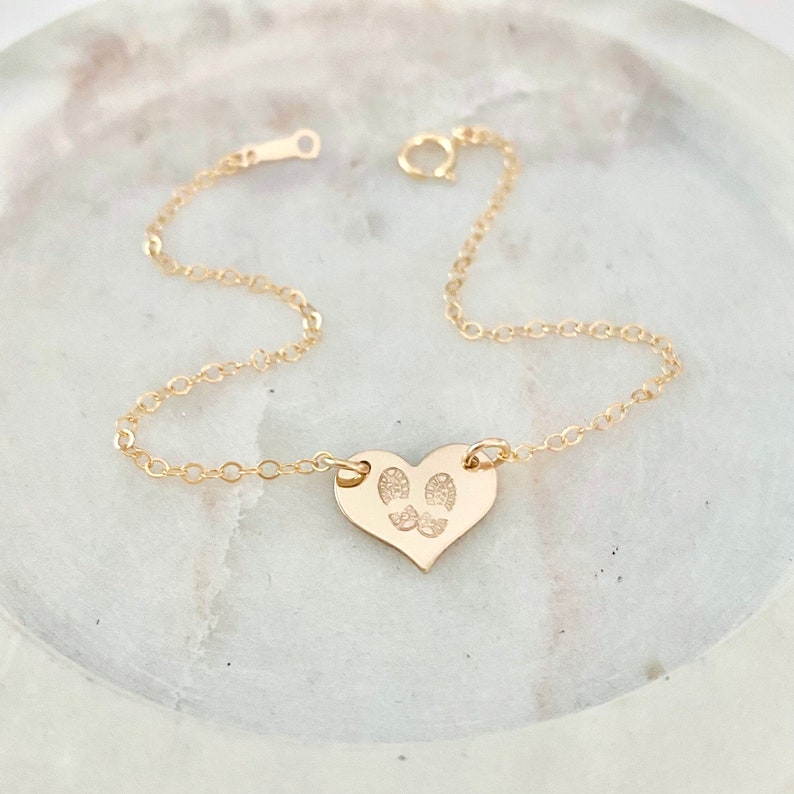 May include: A gold heart-shaped charm bracelet with a delicate chain. The charm features an engraved design of two footprints.