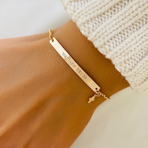 May include: Gold-coloured bar bracelet with the text "MATTHEW 28:20" engraved on it. A small cross charm hangs from the bottom of the bar. The bracelet is on a gold chain.