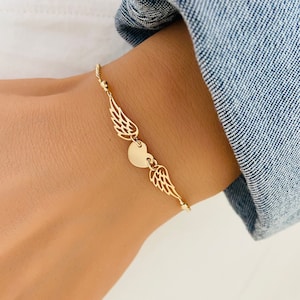 May include: A gold bracelet featuring a heart charm and angel wings. The bracelet is worn on a person's wrist.