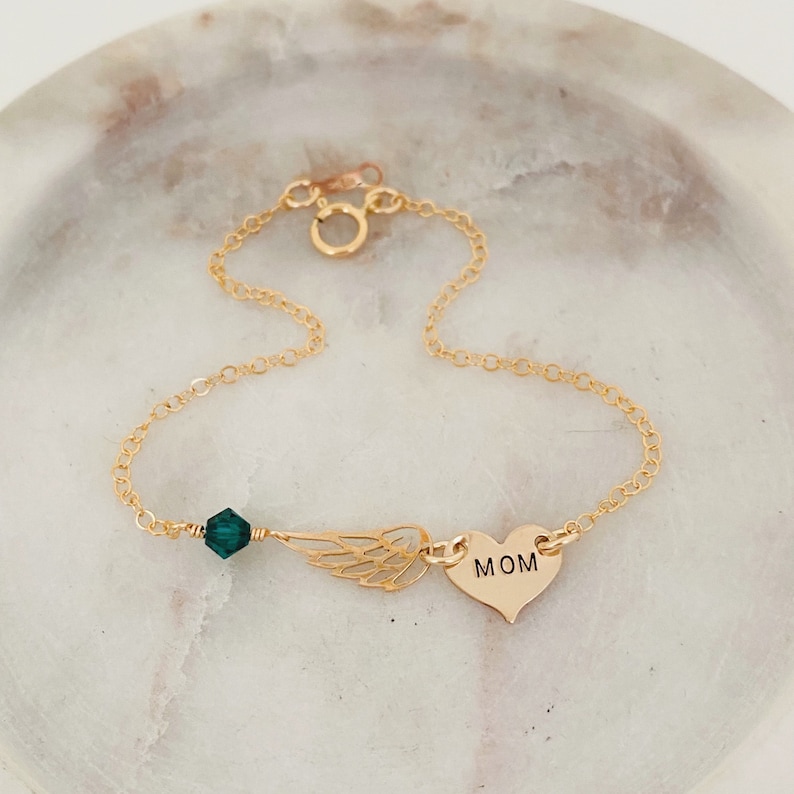 K&ouml;nnte beinhalten: Ein zartes, goldfarbenes Armband mit einem kleinen Herzanh&auml;nger mit der Gravur "MOM". Das Armband enth&auml;lt au&szlig;erdem einen filigranen Fl&uuml;gelanh&auml;nger, eine dunkelgr&uuml;ne Kristallperle und eine Kette mit einem Karabinerverschluss.