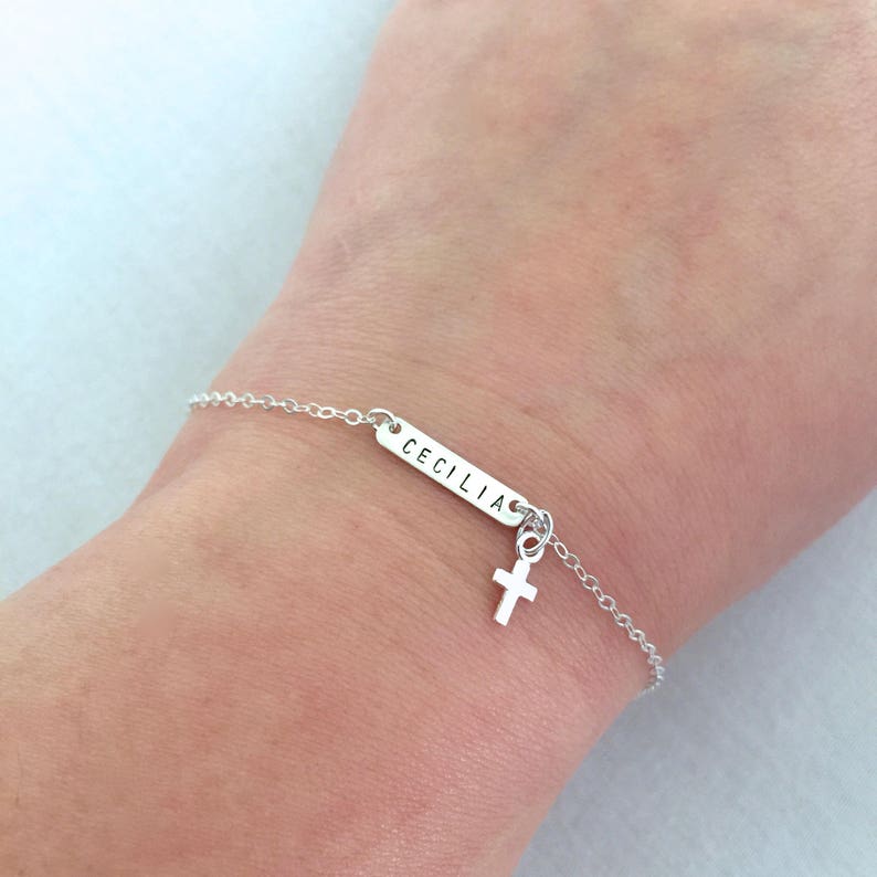 May include: Silver chain bracelet with a small cross charm and a rectangular tag with the name "CECILIA" engraved on it.