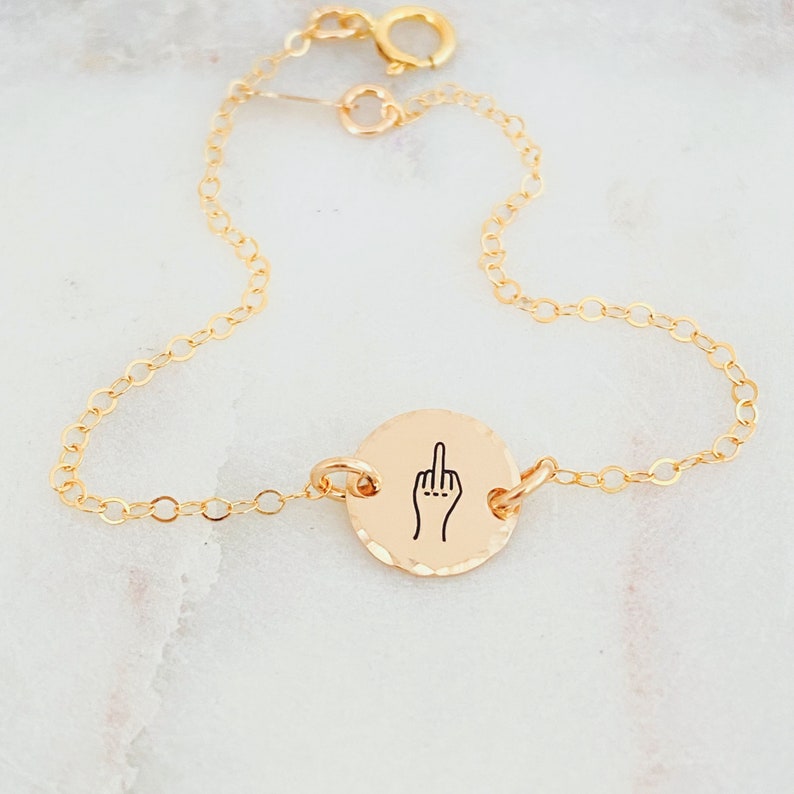 Puede incluir: Pulsera de cadena dorada con un dije redondo que muestra un gesto de dedo medio.