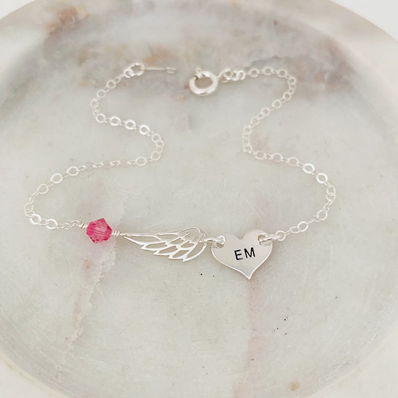 K&ouml;nnte beinhalten: Ein filigranes Silberarmband mit einem Herzanh&auml;nger mit der Gravur "EM", einem ausgeschnittenen Fl&uuml;gel und einer rosa Kristallperle. Das Armband ist durch eine filigrane Kette verbunden und hat einen Verschluss.
