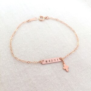 May include: A rose gold chain bracelet with a small cross charm and a rectangular tag engraved with the name "NAOMI".