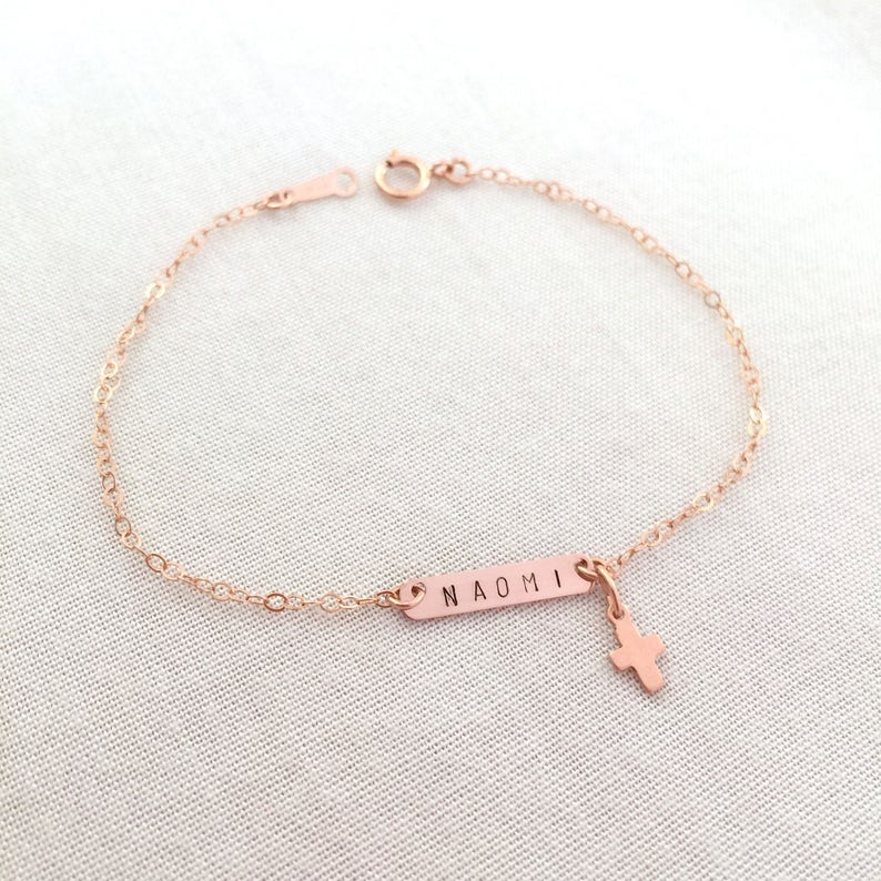 May include: A rose gold chain bracelet with a small cross charm and a rectangular tag engraved with the name "NAOMI".