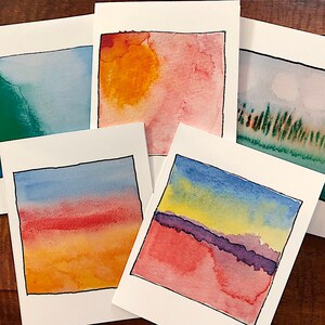 May include: Five watercolor paintings on white card stock, each depicting a different landscape scene. The scenes include a sunset with orange, red, and yellow hues, a blue sky with a yellow sun, a green field with trees, a blue sky with a yellow and purple horizon, and a blue sky with a red and orange horizon.