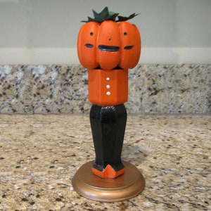 May include: A carved wooden Halloween decoration featuring a three-faced pumpkin head with green leaves. The pumpkin sits atop an orange body with white buttons, black pants, and a gold base. A festive fall accent.