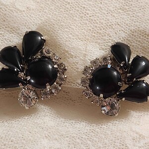 May include: A pair of clip-on earrings featuring a central round black cabochon surrounded by a cluster of clear faceted rhinestones and three black teardrop-shaped stones arranged in a fan-like pattern, all set in a silver-toned metal frame.