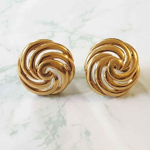 Puede incluir: Un par de pendientes redondos dorados con clip. Cada pendiente presenta un diseño en espiral. Los pendientes están colocados sobre un fondo blanco y verde con vetas de mármol.
