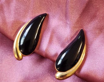 May include: Pair of black teardrop-shaped earrings with gold-colored trim. The earrings have a glossy finish and are set against a pink fabric background. The earrings are a vintage style.