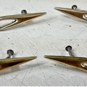 May include: A set of four gold-toned metal drawer pulls, each featuring a sleek, elongated, wing-like shape with a central open cutout design. Each handle is equipped with two small metal mounting screws extending from the back side.