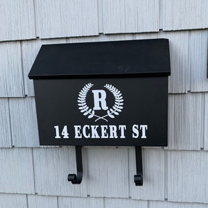 Puede incluir: Buzón negro con una corona blanca y la letra "R" en el centro. El buzón también tiene la dirección "14 ECKERT ST" impresa en blanco. Un timbre negro con un logotipo "ring" blanco está montado a la derecha del buzón.