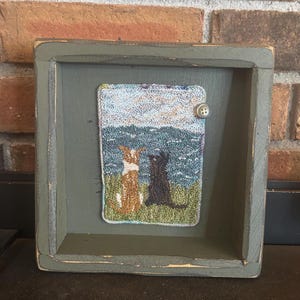 May include: A framed needlepoint artwork depicting two dogs looking out at a blue ocean with a cloudy sky. The dogs are brown and black, and the artwork is framed in a dark green wooden frame.