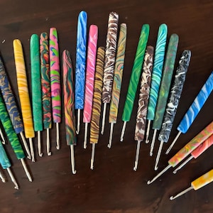 May include: A collection of colorful crochet hooks with swirled and marbled patterns in various colors, including blue, green, yellow, pink, and brown. The hooks are arranged on a dark wooden surface.