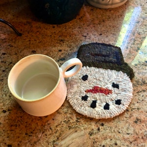 May include: A white ceramic mug with a brown handle sits on a white snowman-shaped crocheted coaster. The snowman has a black hat, black eyes, a red nose, and a black mouth.