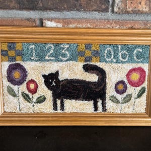 May include: A framed, hand-stitched artwork featuring a black cat, flowers, and the numbers "123" and letters "abc". The cat is centered, with flowers on either side. The background is off-white, with a light brown wooden frame.