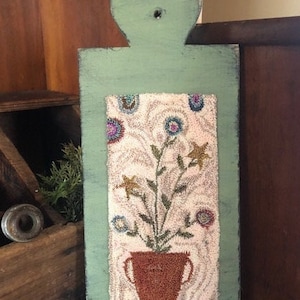 May include: A rustic, rectangular, green wooden wall hanging with a floral design. The central panel features a textured, off-white background with a brown pot holding a floral arrangement with blue, yellow, and purple flowers. The piece has a distressed, vintage aesthetic.