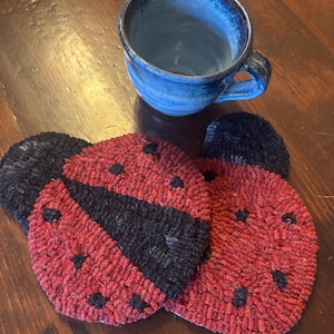 May include: Handmade ladybug-themed coasters. The coasters are red with black accents, including a black stripe down the center and black dots. A blue ceramic mug with a handle sits in the background. The coasters are on a wooden surface.