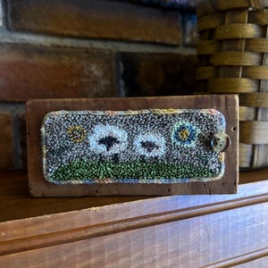 May include: A rectangular wooden box with a hand-stitched wool design featuring two white sheep, a yellow sun, and a blue flower. The box is adorned with a button closure and sits on a wooden shelf.