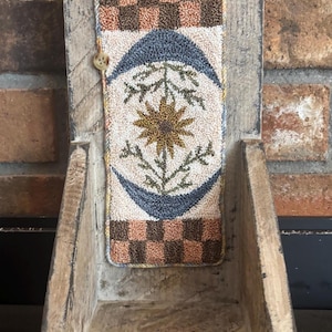 May include: A rustic wooden wall pocket with a decorative, rectangular insert. The insert features a floral design with a sunflower in the center, surrounded by blue crescent shapes and a checkered pattern in brown and tan.