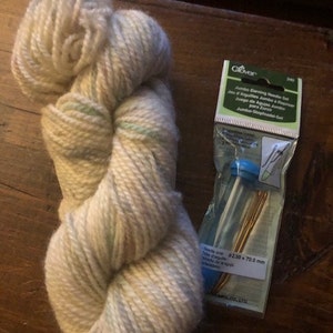 May include: A skein of cream-colored yarn with green and pink flecks, next to a package of Clover jumbo darning needles. The package says "Jumbo Darning Needles Set" and "$2.50 x 70.0 mm".
