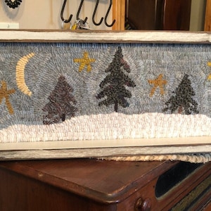 May include: A framed needlepoint wall hanging with a winter scene. The scene features a crescent moon, stars, and trees in a snowy landscape. The frame is white with a distressed finish.
