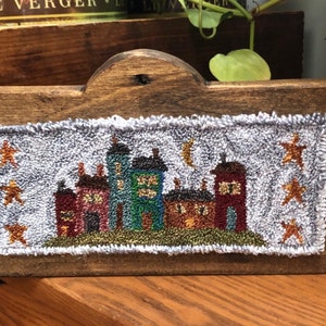 May include: A hand-stitched needlepoint wall hanging featuring a row of colorful houses with a crescent moon and stars against a light blue background. The houses are stitched in shades of red, green, brown, and yellow. The piece is mounted on a wooden frame.