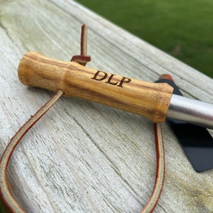 May include: A wooden grilling tool with a bamboo handle, engraved with "DLP". The handle is attached to a metal blade. A brown leather loop is attached to the handle.