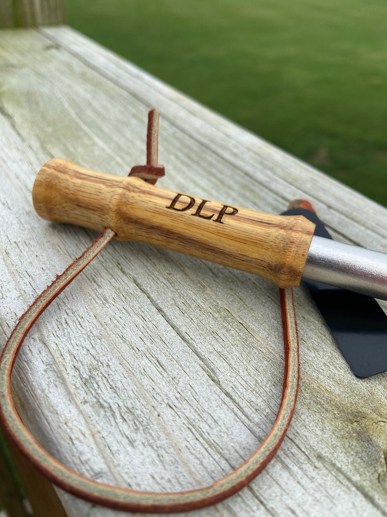 May include: A wooden grilling tool with a bamboo handle, engraved with "DLP". The handle is attached to a metal blade. A brown leather loop is attached to the handle.