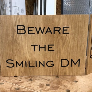 May include: A wooden game master screen with a black and white graphic of a twenty-sided die and the text "Beware the Smiling DM".