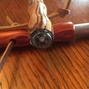 May include: A wooden fire starter with a compass. The handle is made of striped wood and features a small compass at the end. A leather strap is attached for carrying. The fire starter is designed for outdoor use.
