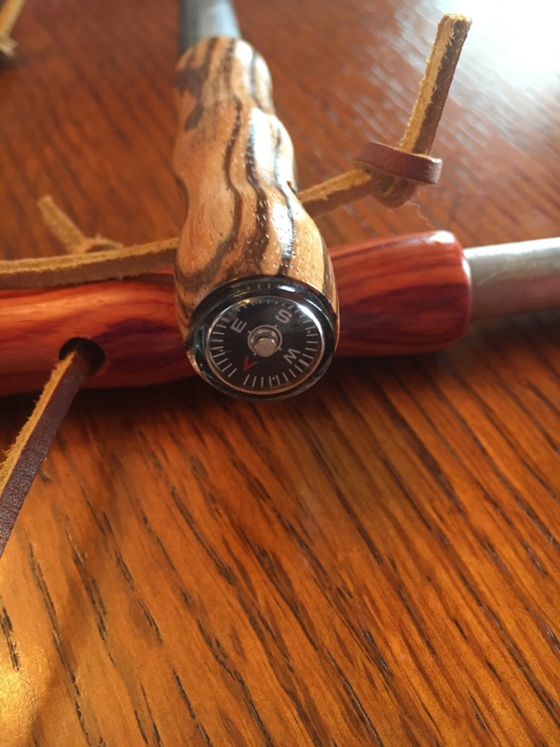 May include: A wooden fire starter with a compass. The handle is made of striped wood and features a small compass at the end. A leather strap is attached for carrying. The fire starter is designed for outdoor use.